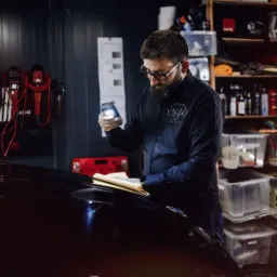 Mężczyzna w okularach z brodą, w ciemnej koszuli z logo, oświetla smartfonem czarny lakierowany samochód, trzymając w ręku dokumenty, w tle regał z pojemnikami i narzędziami.