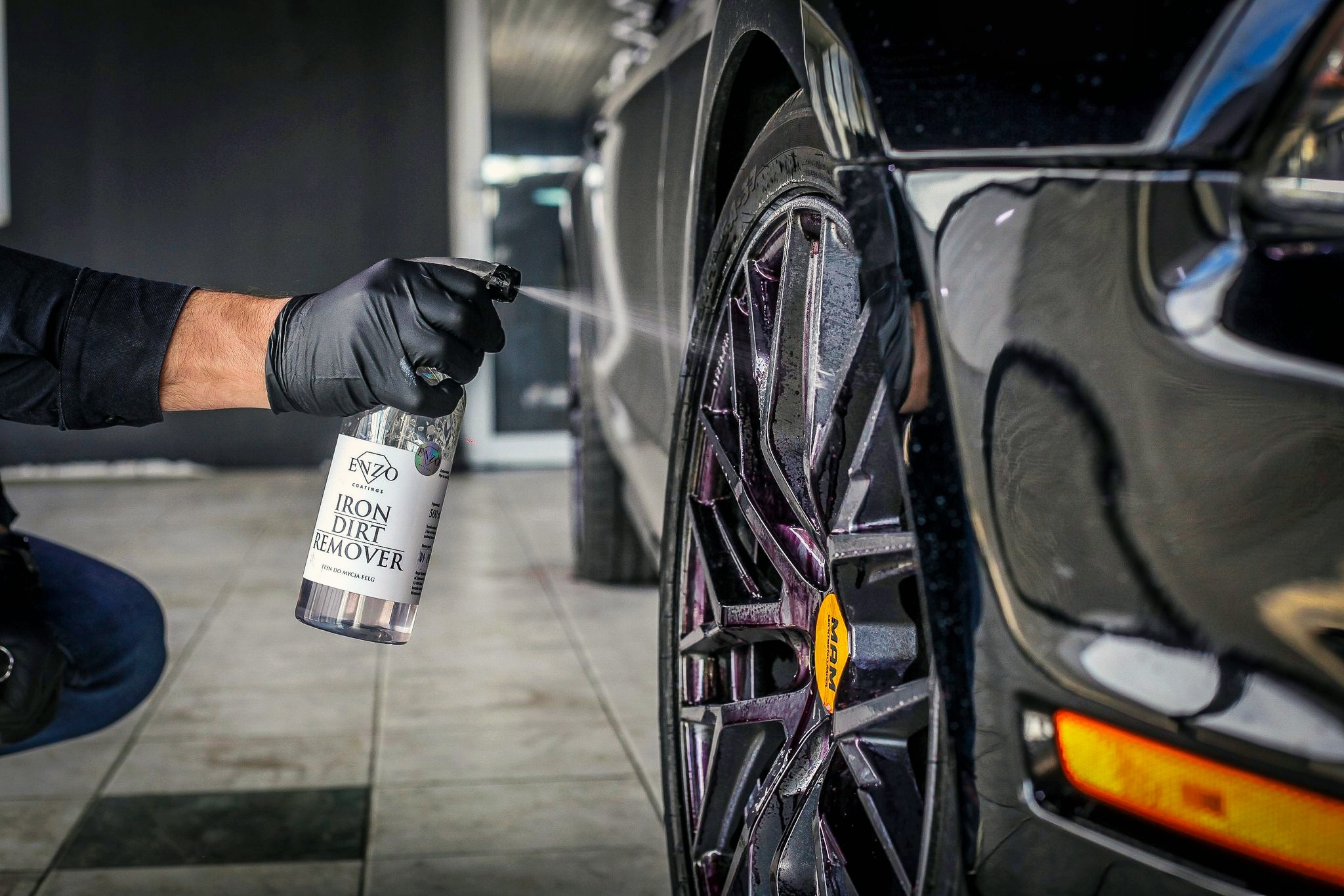 Czyszczenie felgi aluminiowej czarnym preparatem do usuwania zabrudzeń metalicznych, widoczna rozpylona ciecz i pędzel w rękawicy ochronnej.