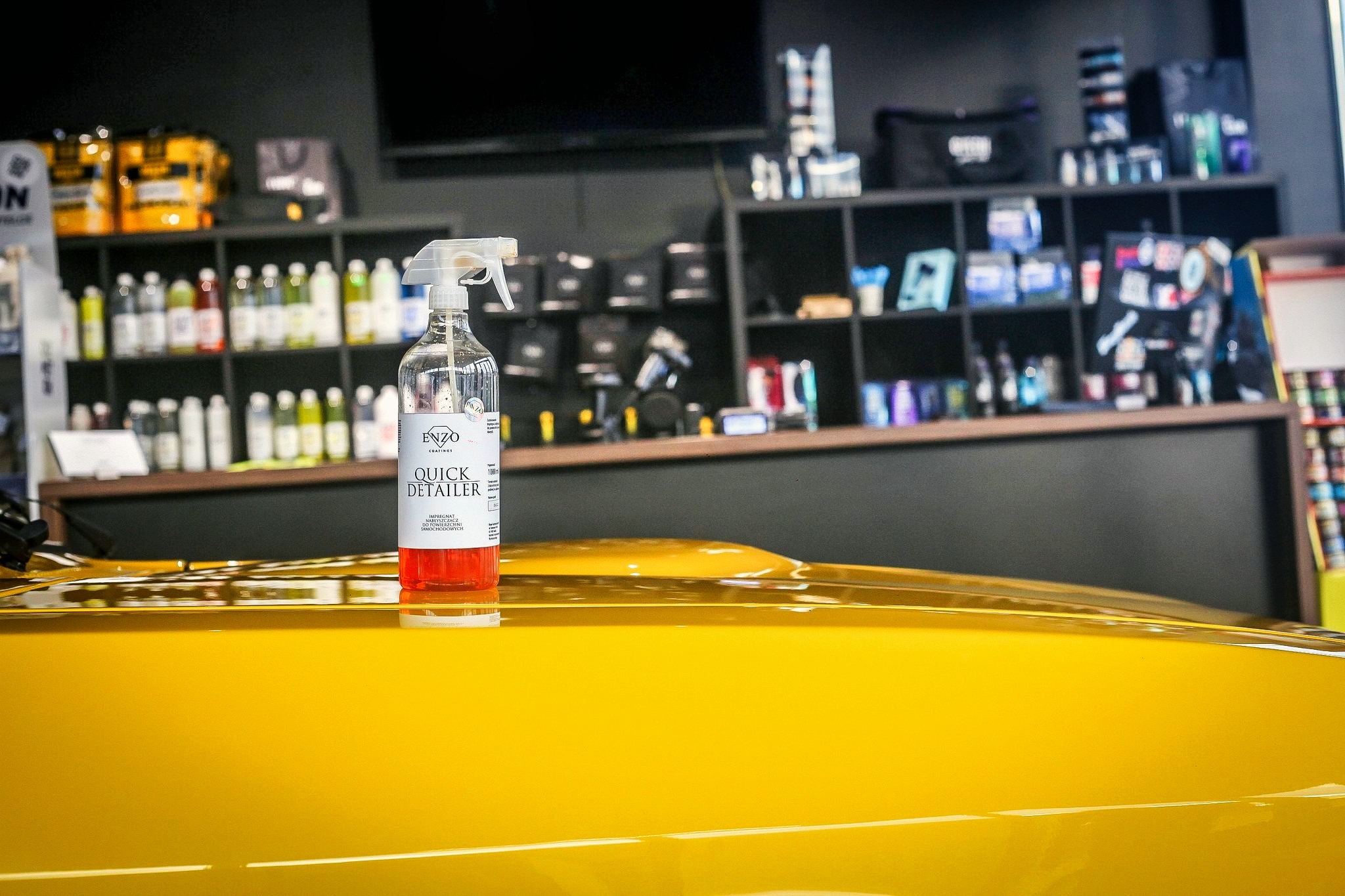 Butelka Quick Detailer Enzo Coatings na żółtym, błyszczącym lakierze samochodu, w tle półki z produktami do detailingu.