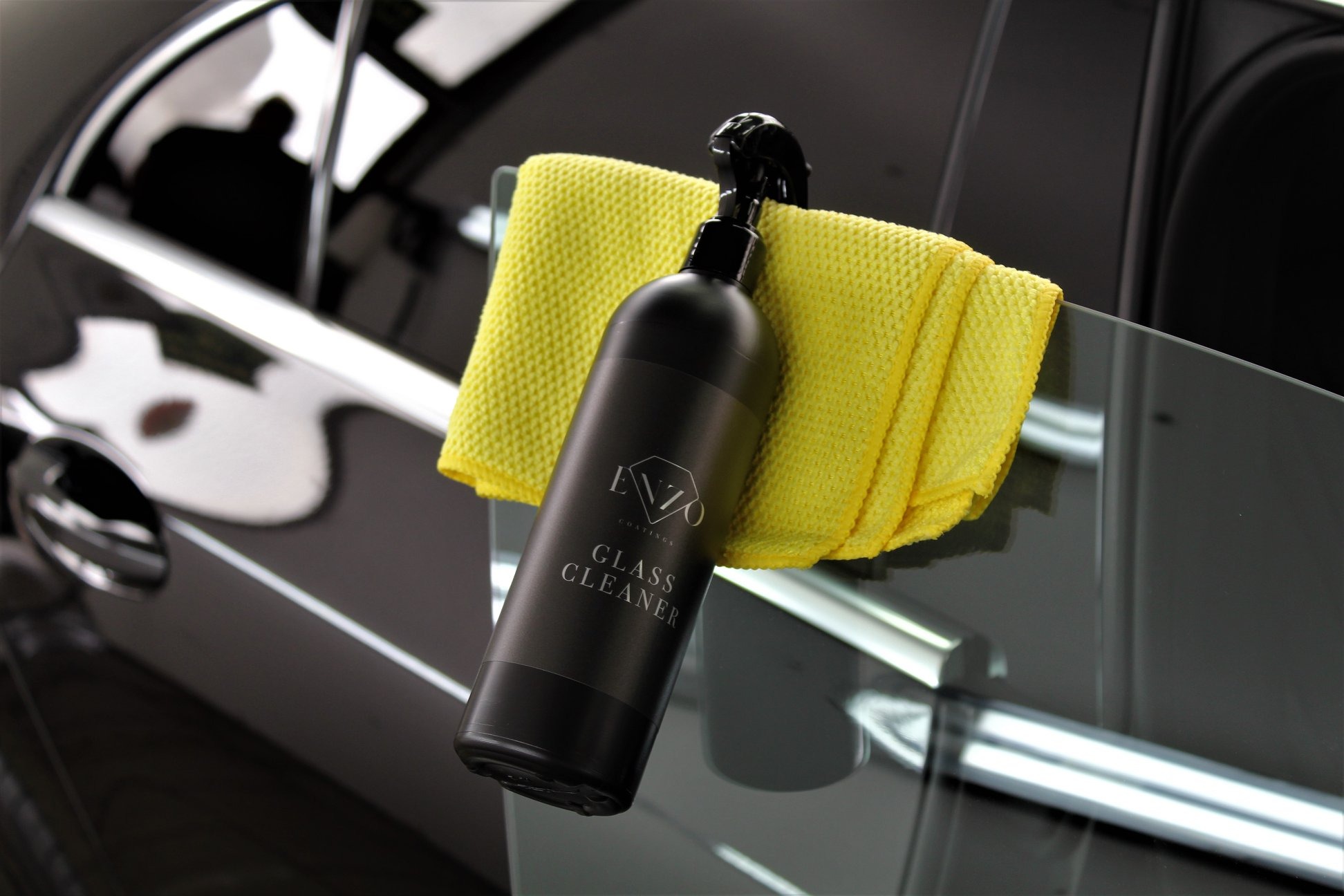 Butelka czarnego płynu do czyszczenia szyb samochodowych marki Enzo leży na żółtej ściereczce z mikrofibry, oparta o szybę czarnego samochodu.