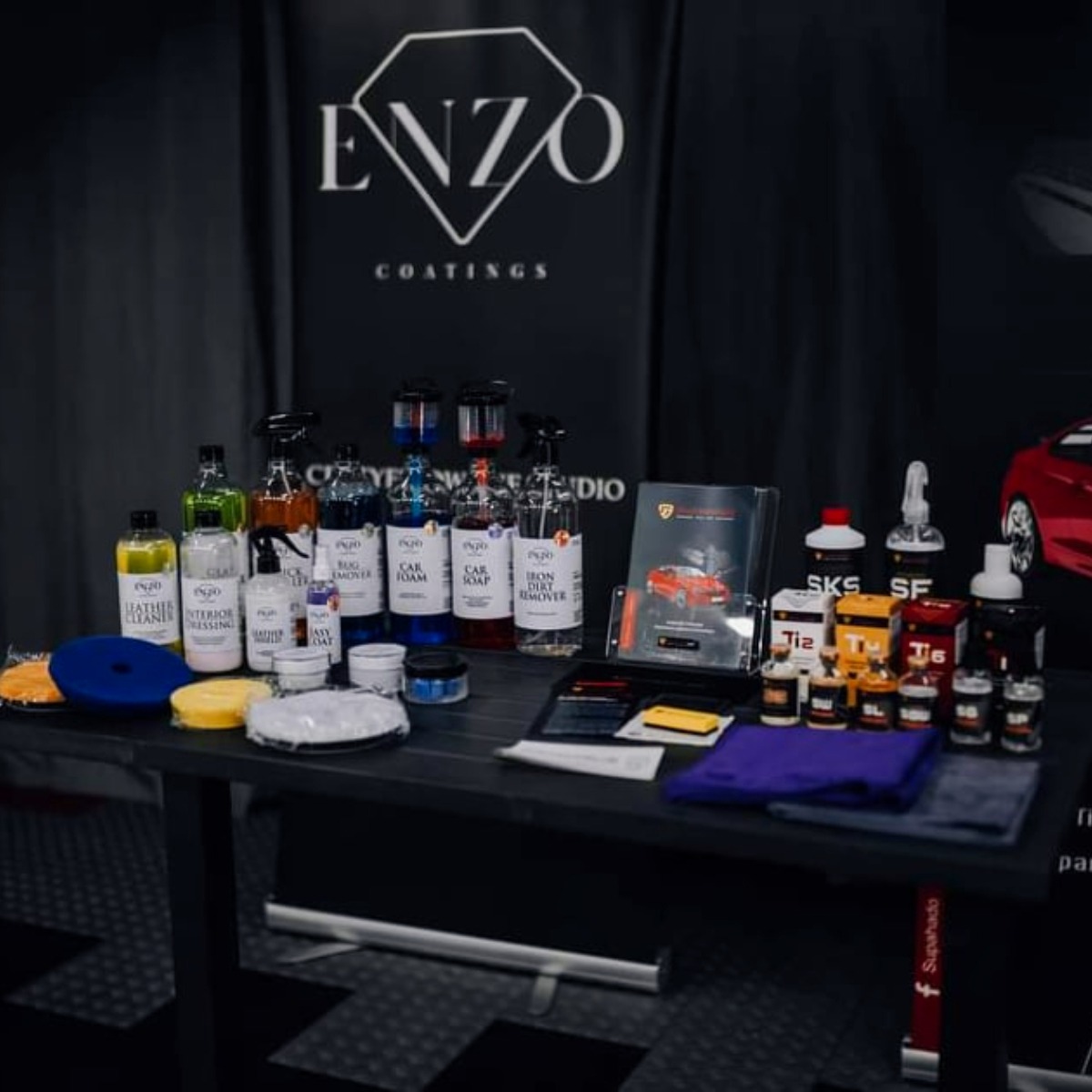 Zestaw środków do pielęgnacji samochodu marki Enzo Coatings, prezentowany na stole z logo firmy w tle, obejmujący preparaty do czyszczenia skóry, usuwania owadów, pianę do mycia, szampon i preparat...