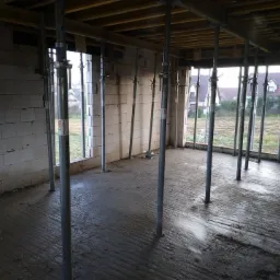 Wnętrze domu w trakcie budowy: murowane ściany z betonu komórkowego, mokra wylewka podłogowa z widocznymi śladami kielni, metalowe stemple podtrzymujące drewniany szalunek stropu, widok na domy...