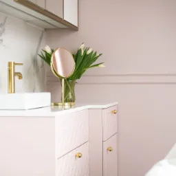 Umywalka nablatowa, złota bateria, lustro na blacie i bukiet białych tulipanów w szklanym wazonie na tle pudrowo-różowej ściany z listwami.
