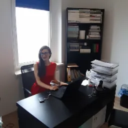 Kobieta w czerwonej sukience siedzi przy czarnym biurku z laptopem w jasnym biurze, za nią regał z książkami i dokumentami.