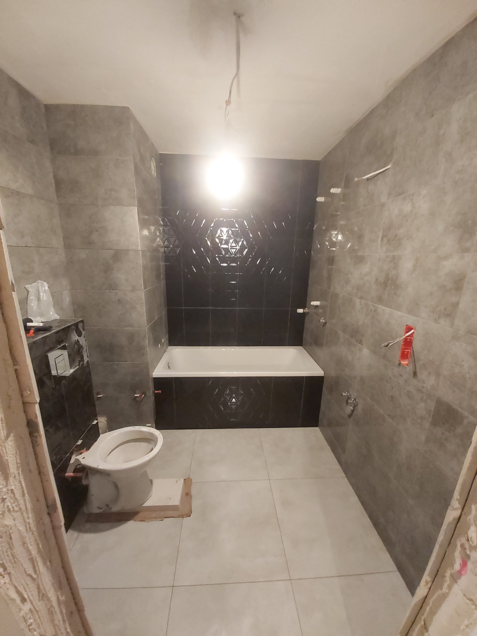 Wnętrze łazienki w trakcie remontu: szare płytki imitujące kamień na ścianach i podłodze, czarna ściana nad wanną z geometrycznym wzorem, częściowo zamontowana toaleta na podeście, widoczne...