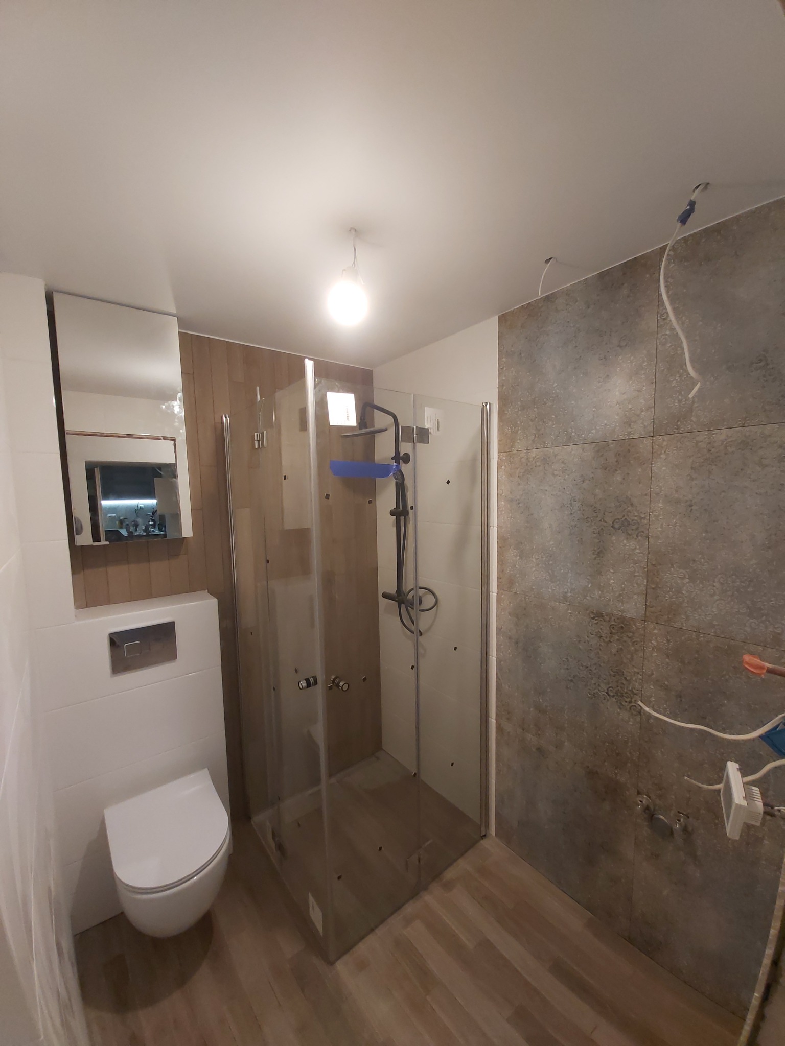 Nowoczesna łazienka w trakcie remontu: kabina prysznicowa ze szklanymi drzwiami, wisząca toaleta z chromowanym przyciskiem, lustro z oświetleniem, szare płytki imitujące kamień na ścianie...