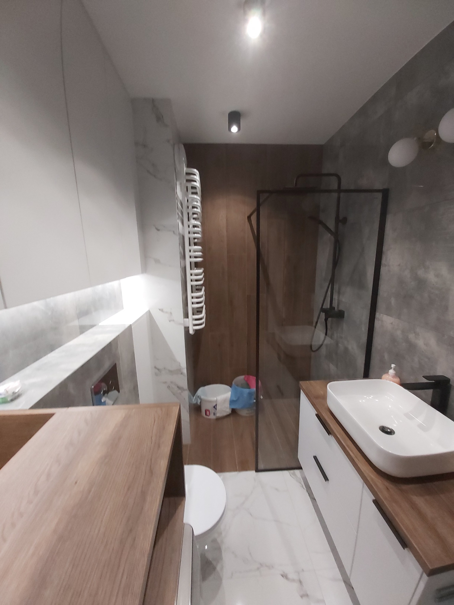 Nowoczesna łazienka z kabiną prysznicową w czarnej ramie, umywalką nablatową na szafce z drewnianym blatem, grzejnikiem drabinkowym i marmurową posadzką.