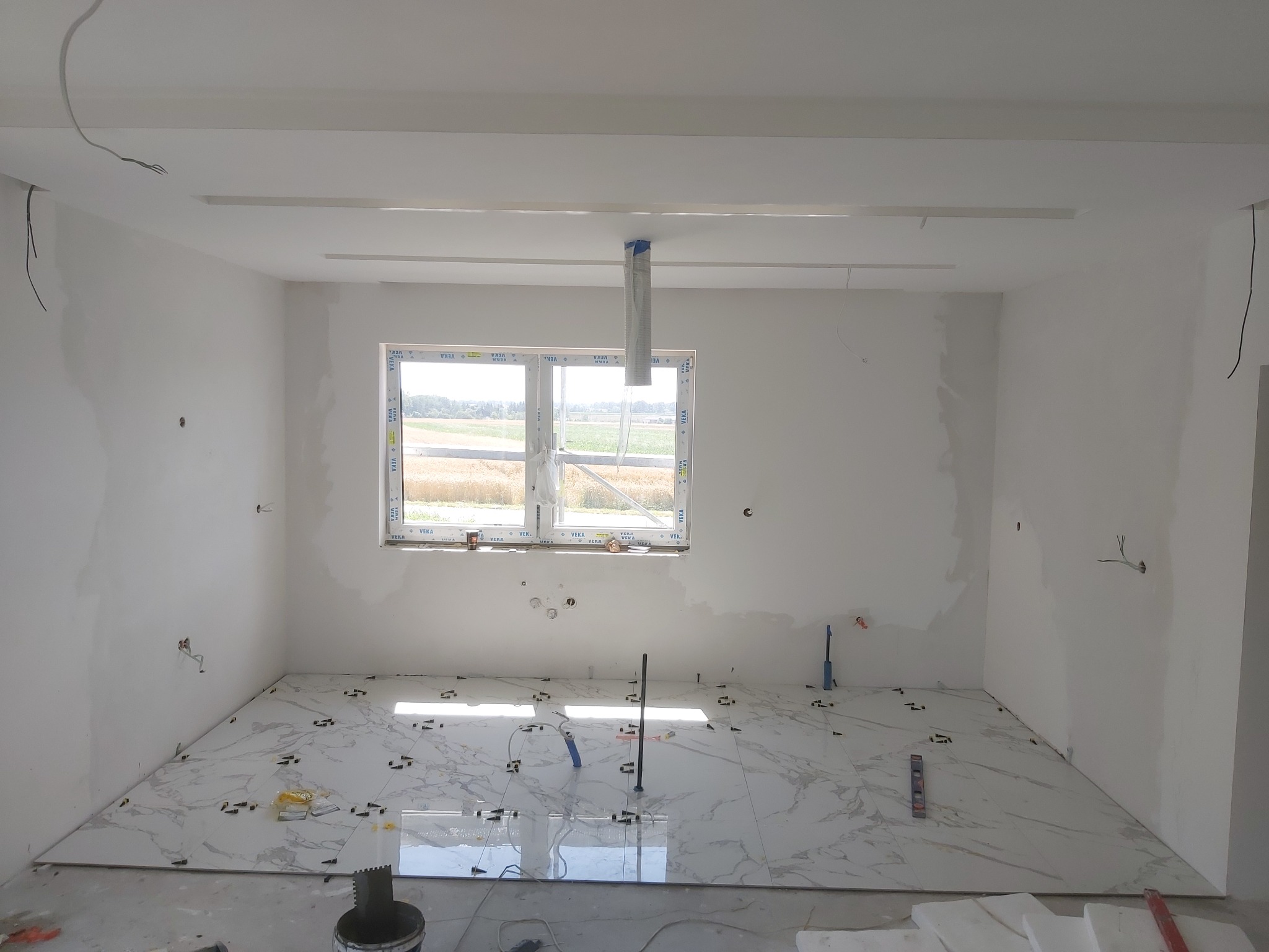 Układanie płytek podłogowych imitujących marmur w jasnym, pustym pokoju z oknem; widoczne kliny poziomujące i narzędzia glazurnicze.