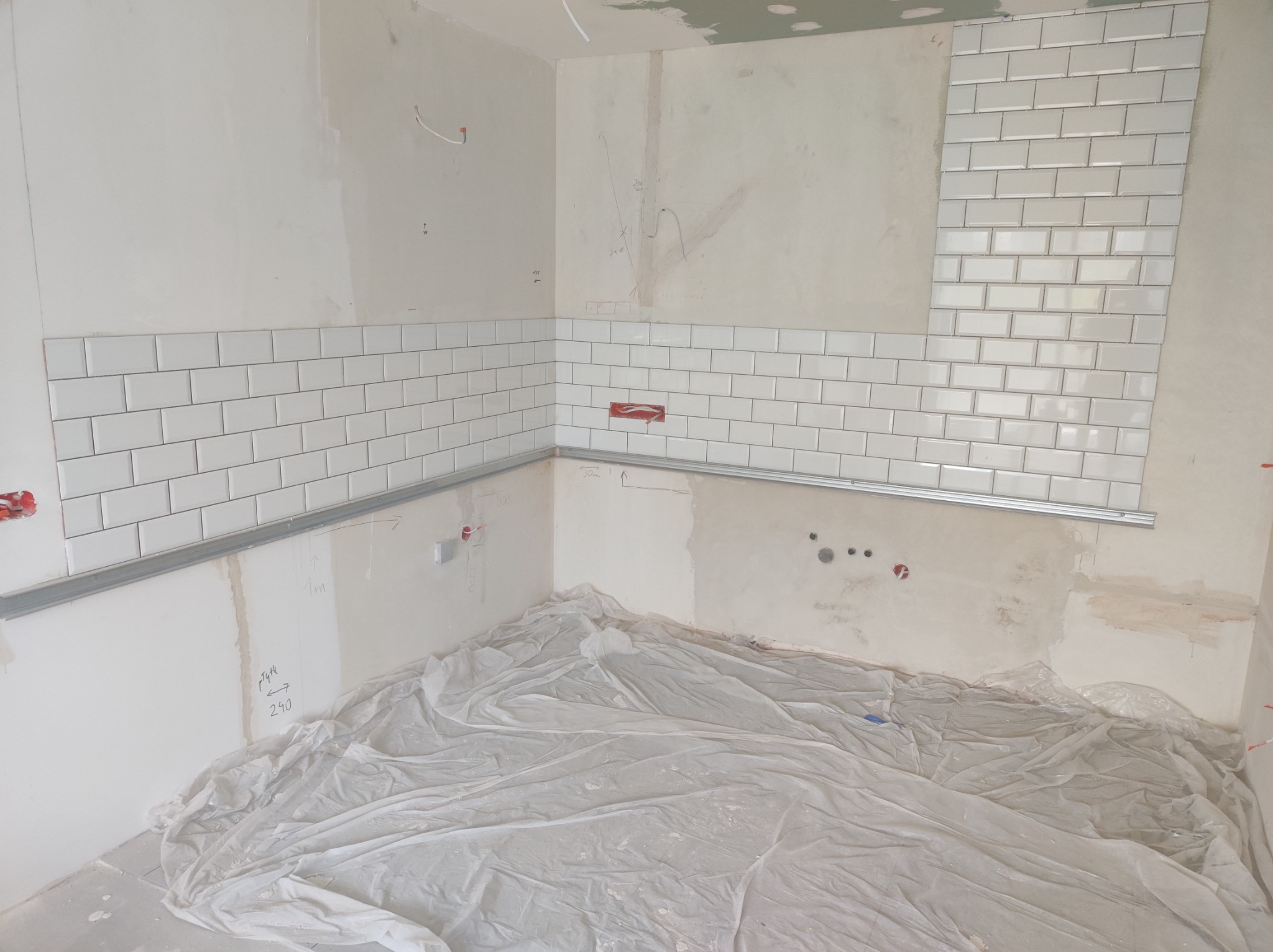 Fragment ściany w trakcie układania białych, prostokątnych płytek typu metro, zabezpieczona folią podłoga, widoczne punkty elektryczne i metalowe listwy montażowe.