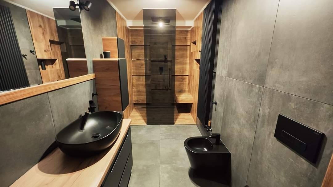 Nowoczesna łazienka z szarymi płytkami, drewnianymi akcentami i czarną armaturą: umywalka nablatowa, bidet i kabina prysznicowa typu walk-in z czarnymi drzwiami.