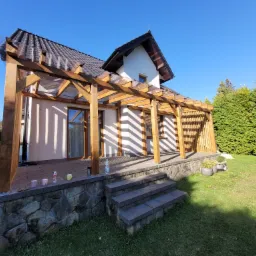 Drewniana pergola z przezroczystym zadaszeniem przylegająca do domu, z kamienną podmurówką i schodami, widok z ogrodu w słoneczny dzień.