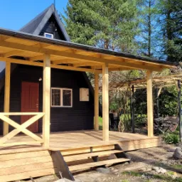 Czarny dom z drewnianą werandą i balustradą, widok z zewnątrz, w tle zielone drzewa i błękitne niebo, na pierwszym planie ziemia i resztki materiałów budowlanych.