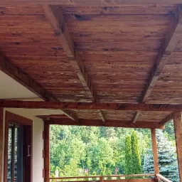 Drewniana konstrukcja zadaszenia tarasu przylegająca do budynku, z widokiem na zielony ogr&oacute;d i las w tle.