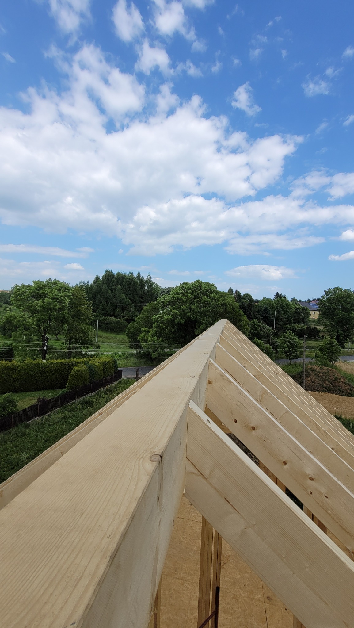 Konstrukcja więźby dachowej z jasnego drewna, widok z góry na tle błękitnego nieba z chmurami i zielonego krajobrazu z drzewami.