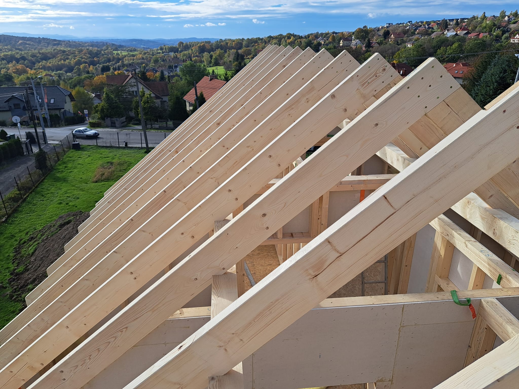 Konstrukcja więźby dachowej z jasnego drewna na tle zielonego krajobrazu z zabudowaniami, widok z góry na wczesnym etapie budowy.