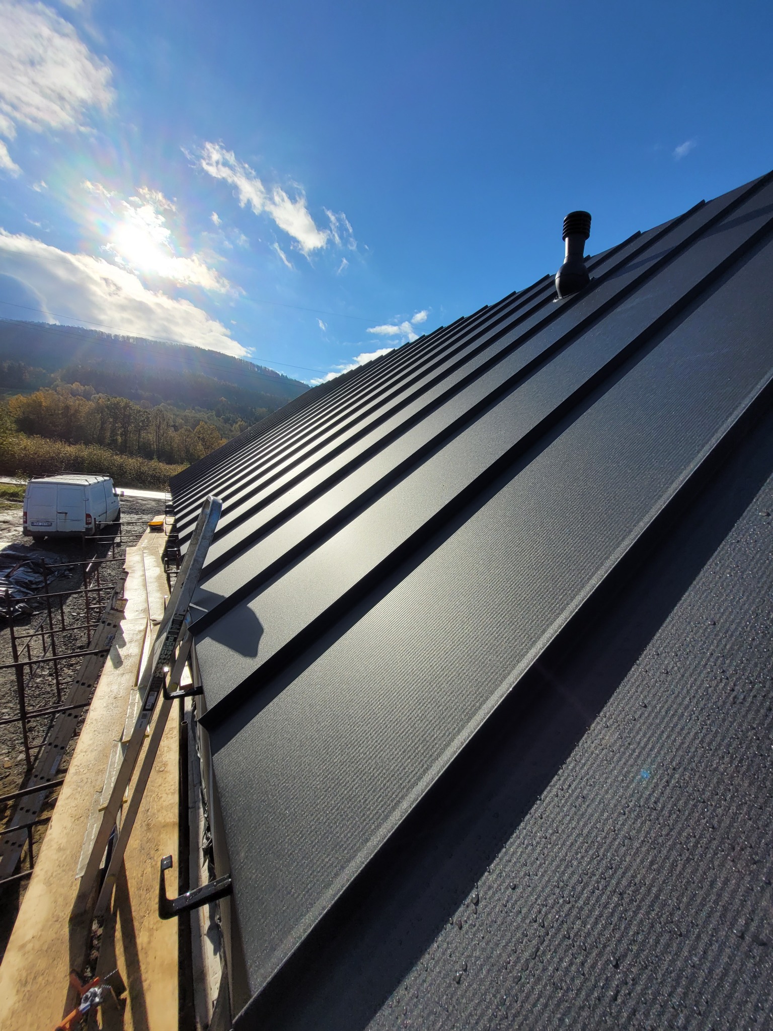 Nowy, czarny dach z blachy na tle błękitnego nieba i zielonych wzgórz, widoczny kominek wentylacyjny i rusztowanie.