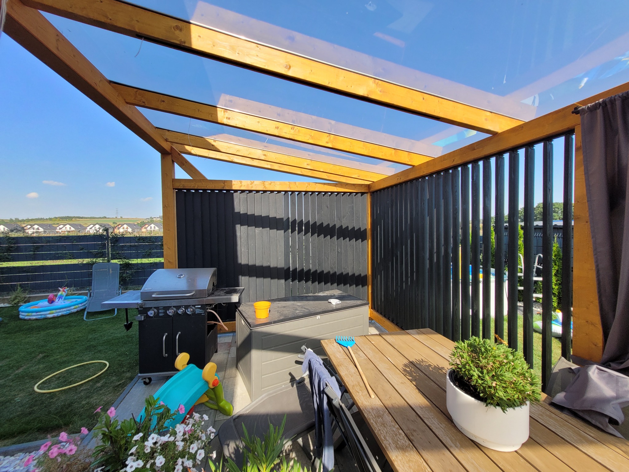 Zadaszona konstrukcja z drewna i czarnych paneli, z widocznym grillem, meblami ogrodowymi i zabawkami na trawniku w tle.