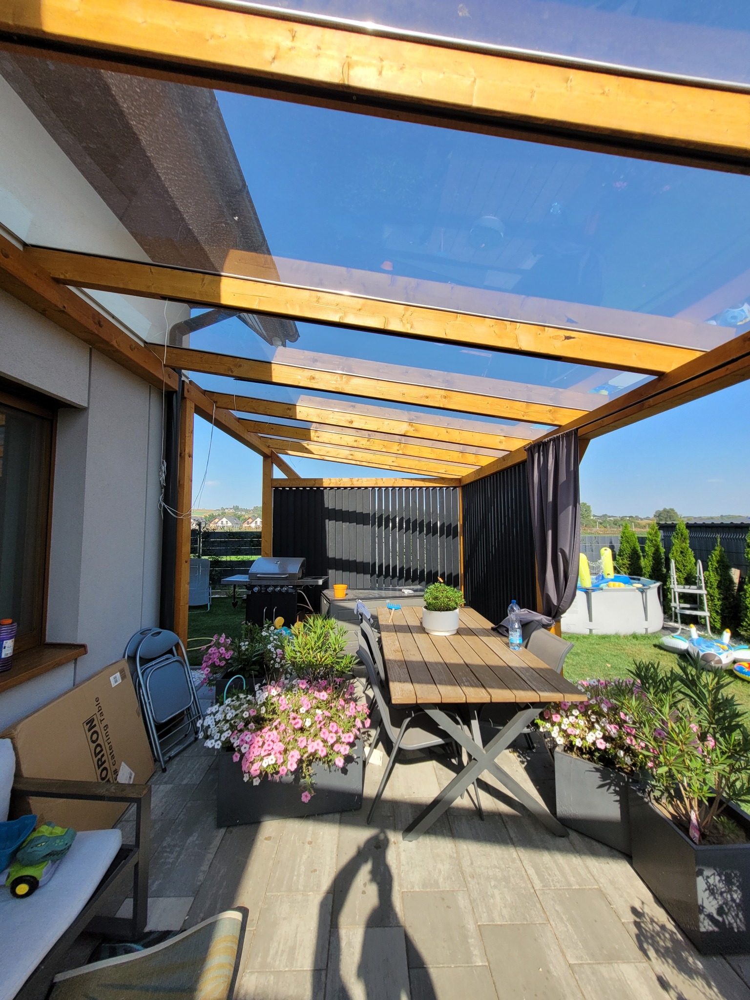 Zadaszone miejsce do wypoczynku na zewnątrz z drewnianą konstrukcją i przezroczystym dachem, stół z krzesłami, kwiaty w donicach, grill i basen w tle.