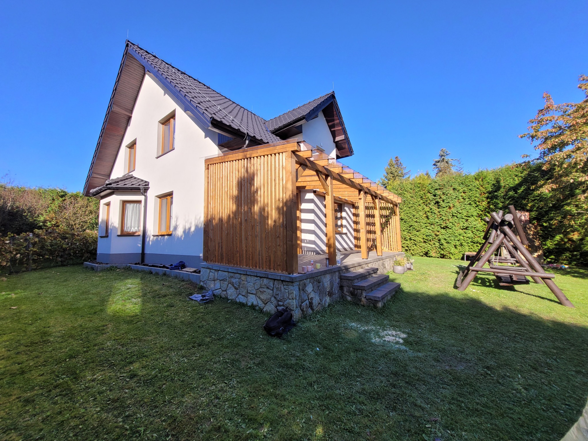 Nowoczesny dom z jasną elewacją i drewnianą pergolą z pionowymi deskami, kamiennym podmurówką i schodami, otoczony zielonym trawnikiem i krzewami w słoneczny dzień.