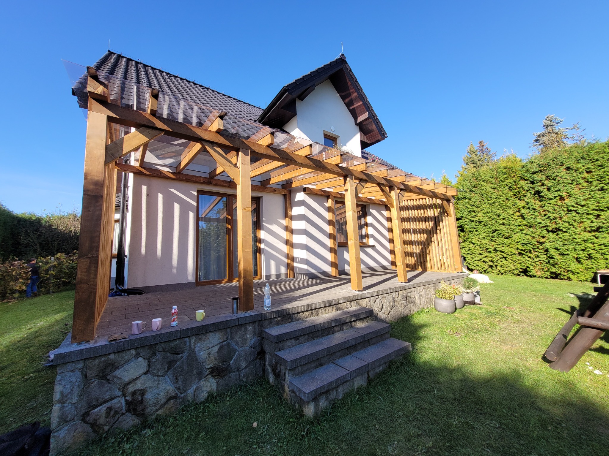 Drewniana pergola z przezroczystym zadaszeniem przylegająca do domu, z kamienną podmurówką i schodami, widok z ogrodu w słoneczny dzień.