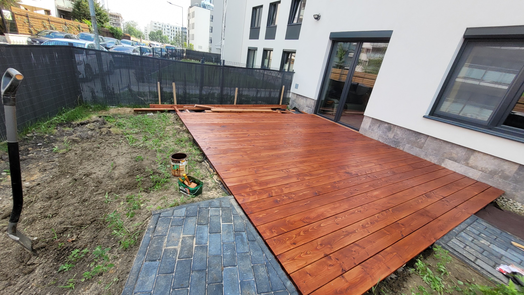 Brązowy taras z desek w trakcie budowy przy nowoczesnym domu, widoczne narzędzia i puszka z impregnatem, teren wokół w trakcie przygotowywania.