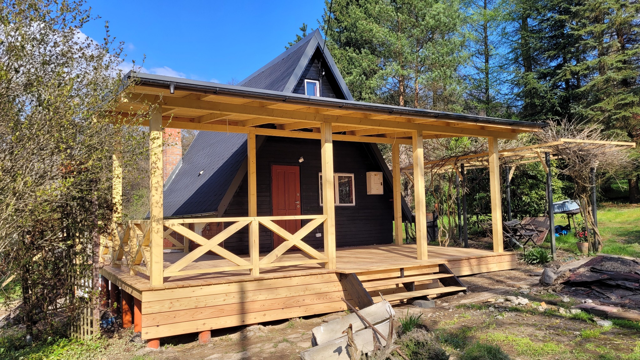 Czarny dom w stylu A-frame z nowym, jasnym drewnianym tarasem i pergolą w ogrodzie, widok z przodu na tle drzew i błękitnego nieba.