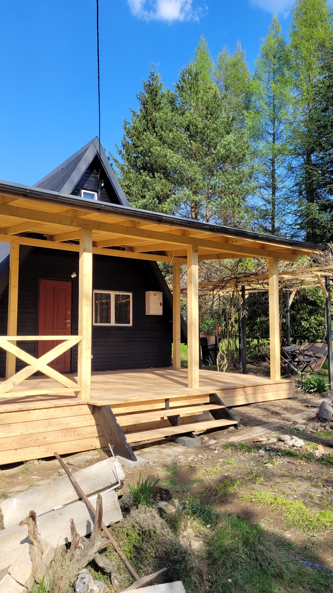 Czarny dom z drewnianą werandą i balustradą, widok z zewnątrz, w tle zielone drzewa i błękitne niebo, na pierwszym planie ziemia i resztki materiałów budowlanych.