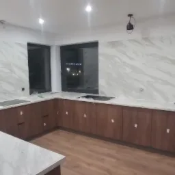 Nowoczesna kuchnia z brązowymi szafkami i blatem imitującym marmur, widoczne dwa okna i wiszące lampy, fragment wyspy kuchennej w folii ochronnej.