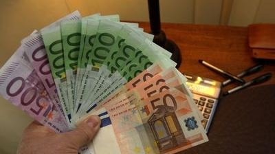 Dłoń trzymająca wachlarz banknotów euro o różnych nominałach (500, 100, 50, 20) na tle biurka z kalkulatorem i długopisami.