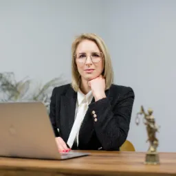 Profesjonalna blondynka w tweedowej marynarce i okularach, siedząca za biurkiem z laptopem, obok statuetki Temidy