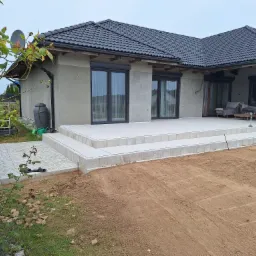 Nowoczesny dom jednorodzinny z szarą elewacją w trakcie wykończenia, z tarasem wyłożonym jasnymi płytami i przygotowanym terenem pod ogród. Widoczne elementy instalacji odgromowej i zbiornik...