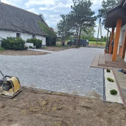 Świeżo ułożona, szara kostka brukowa prowadząca do domu w Barcinie, z widoczną zagęszczarką na pierwszym planie i ozdobnym pasem z białych kamieni i zieleni przy budynku.