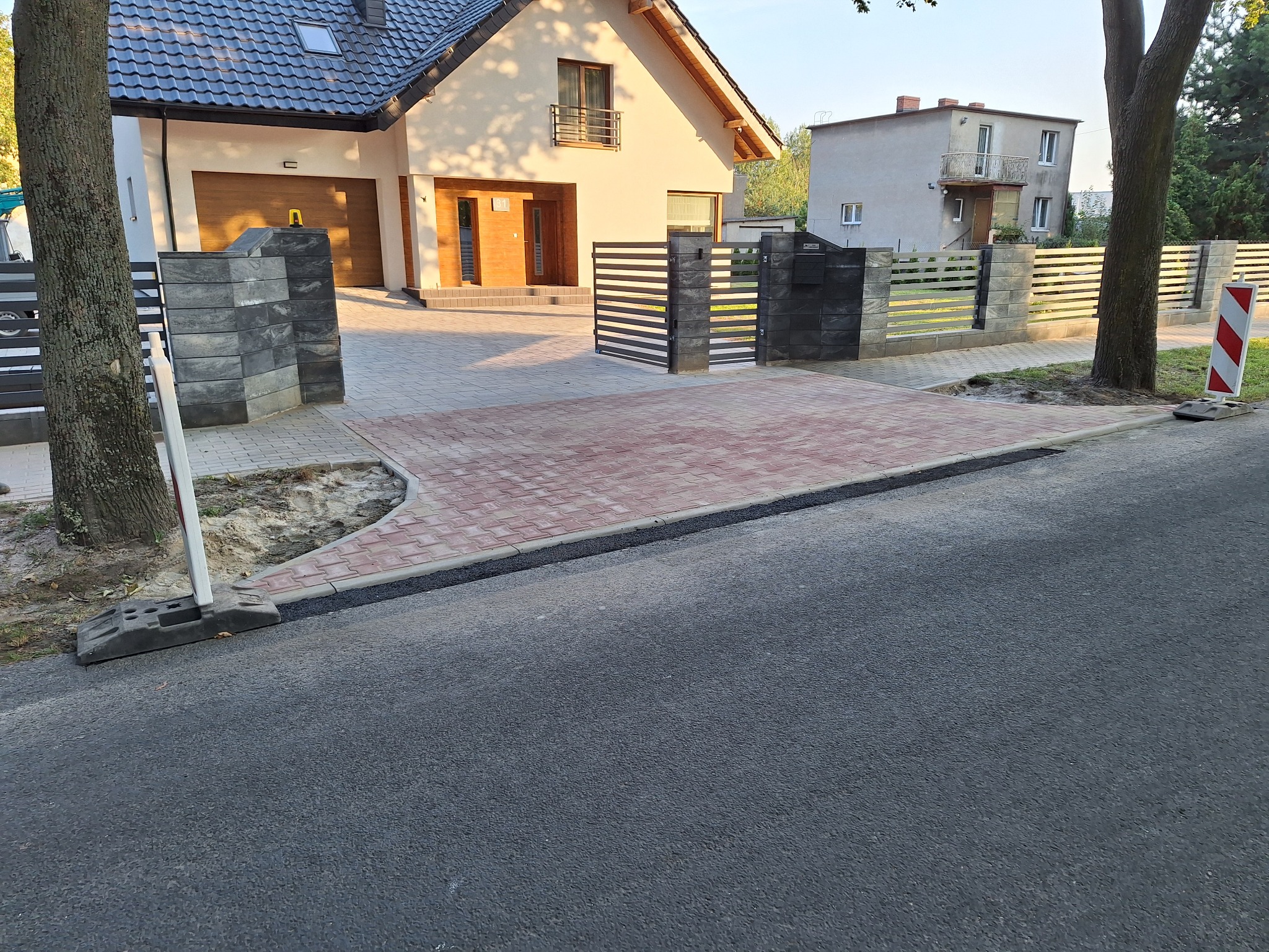 Nowo ułożona, czerwono-szara kostka brukowa na podjeździe przed domem z szarą dachówką, z nowoczesnym ogrodzeniem panelowym i słupkami z szarego betonu łupanego, widok z ulicy.