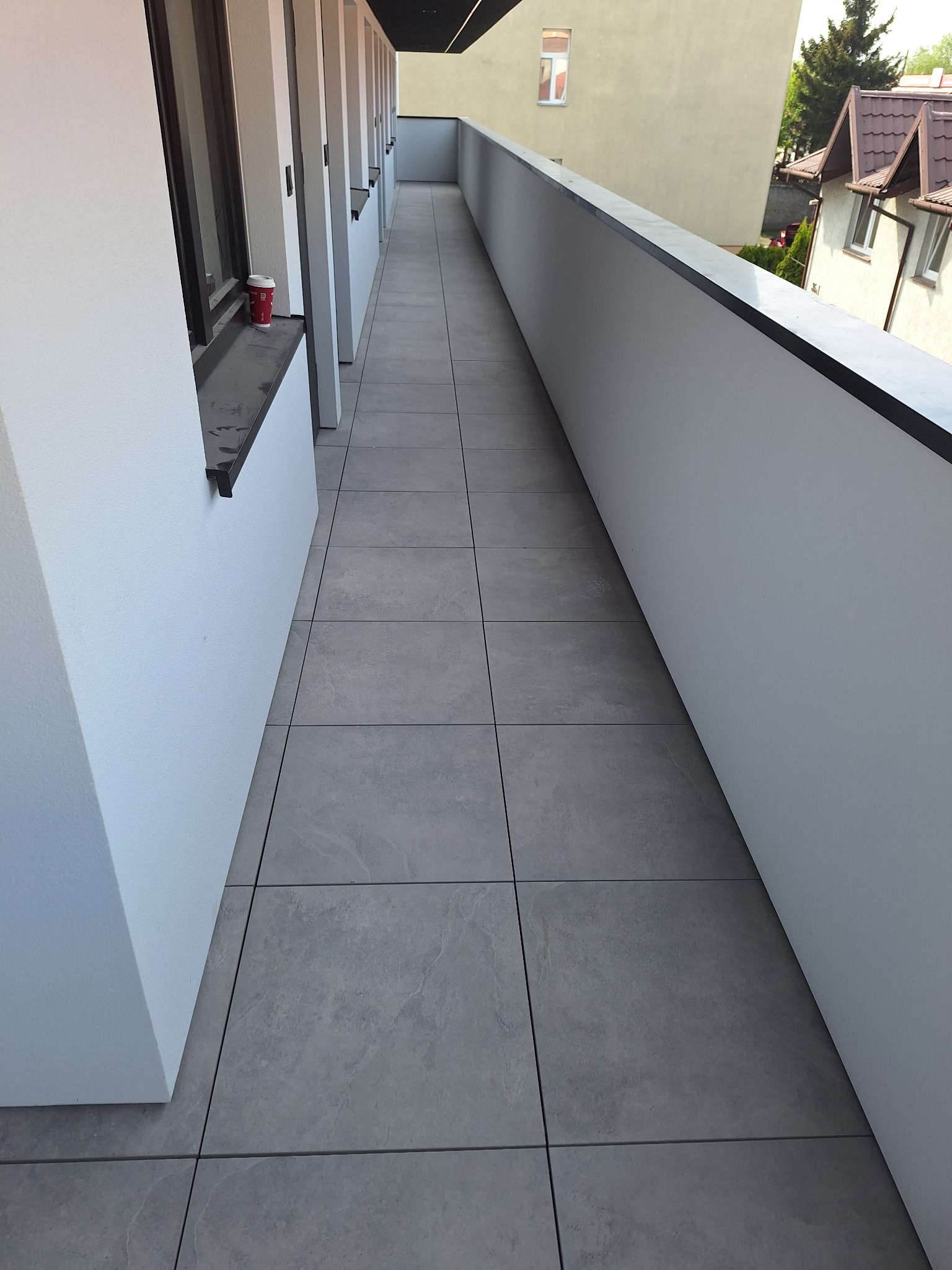 Długi, wąski balkon wyłożony szarymi płytkami, białe ściany z czarnymi akcentami, kubek po kawie na parapecie, widok na budynki i drzewa w tle.
