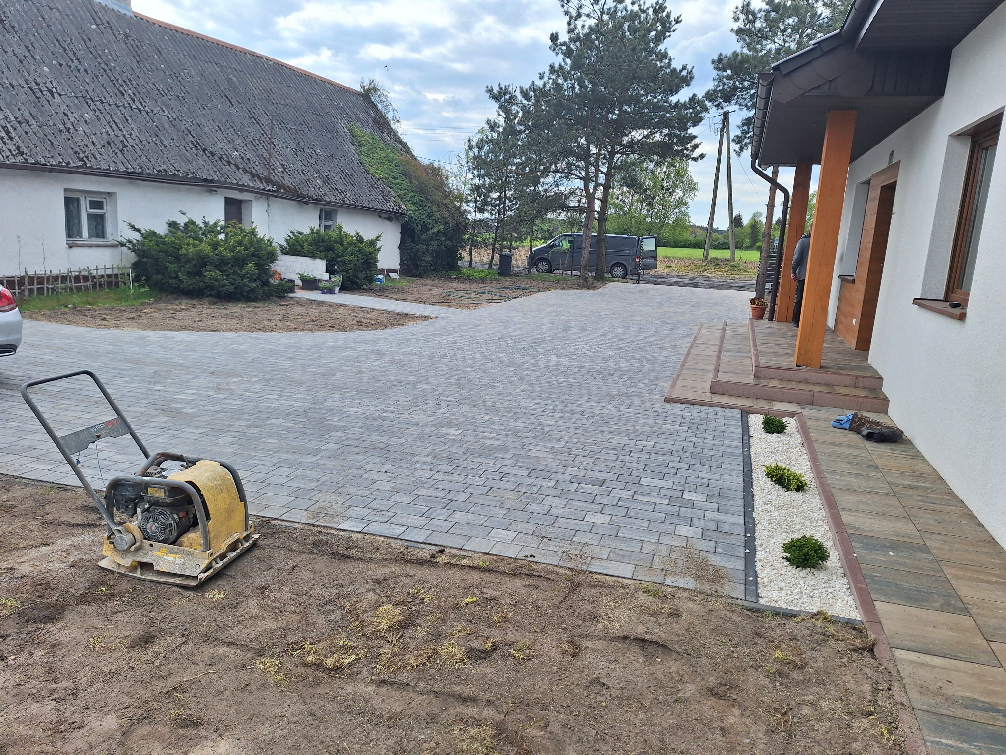 Świeżo ułożona, szara kostka brukowa prowadząca do domu w Barcinie, z widoczną zagęszczarką na pierwszym planie i ozdobnym pasem z białych kamieni i zieleni przy budynku.
