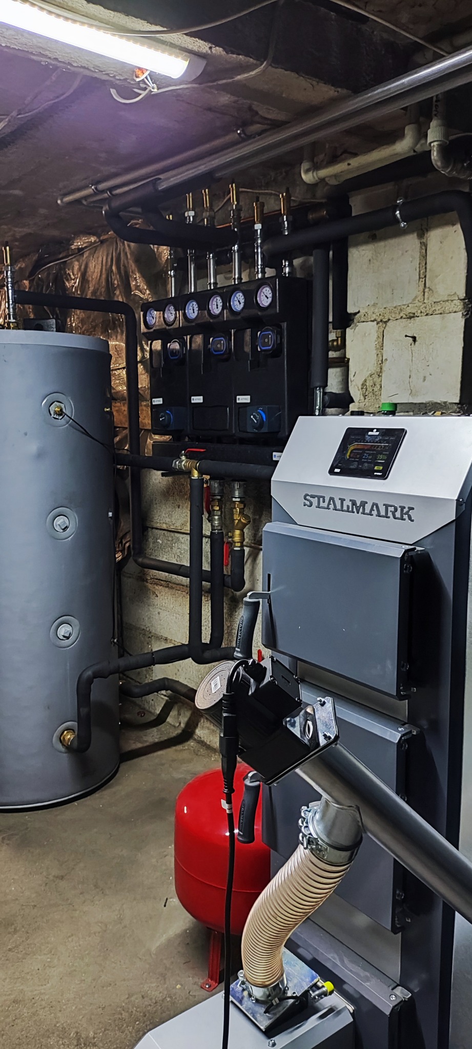 Nowoczesna kotłownia z szarym piecem Stalmark, zbiornikiem akumulacyjnym, czarnymi rurami instalacji grzewczej i czerwonym zbiornikiem wyrównawczym ciśnienia.