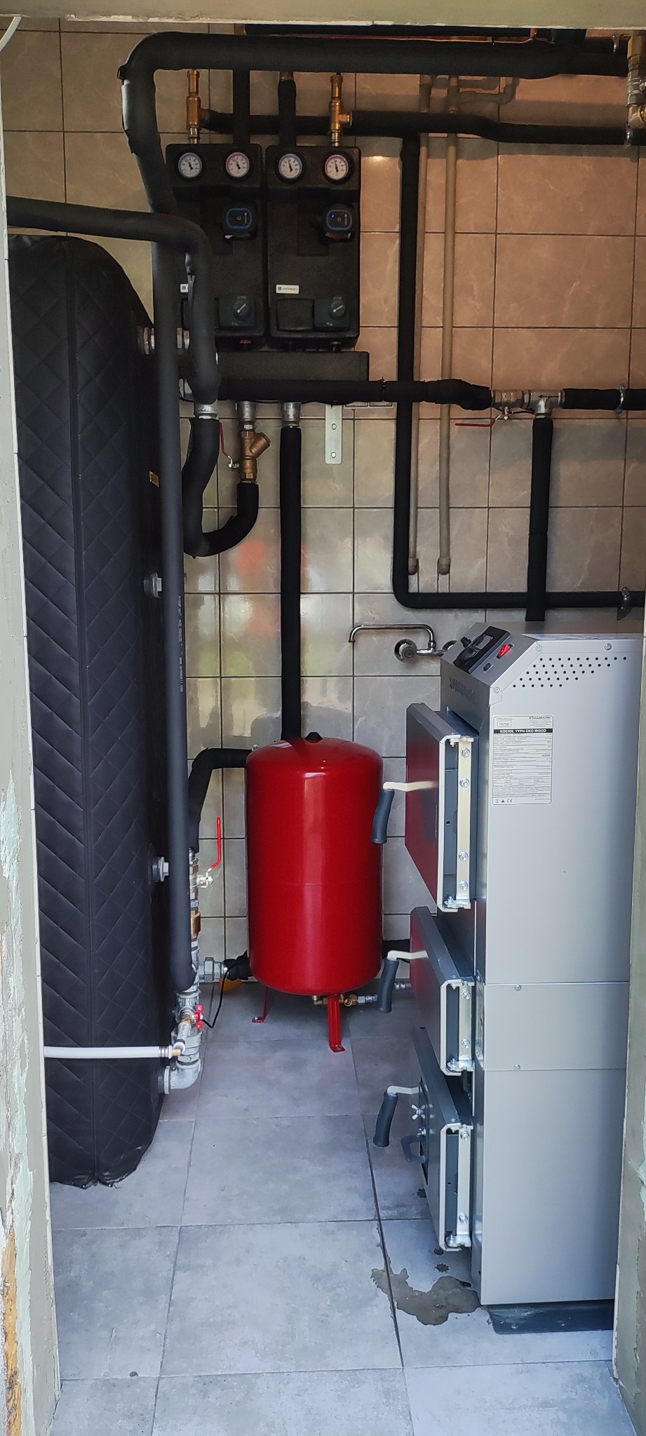 Nowoczesna kotłownia z piecem na paliwo stałe, czerwonym zbiornikiem wyrównawczym i czarnym akumulatorem ciepła, widok na instalację grzewczą z rurami i zaworami.