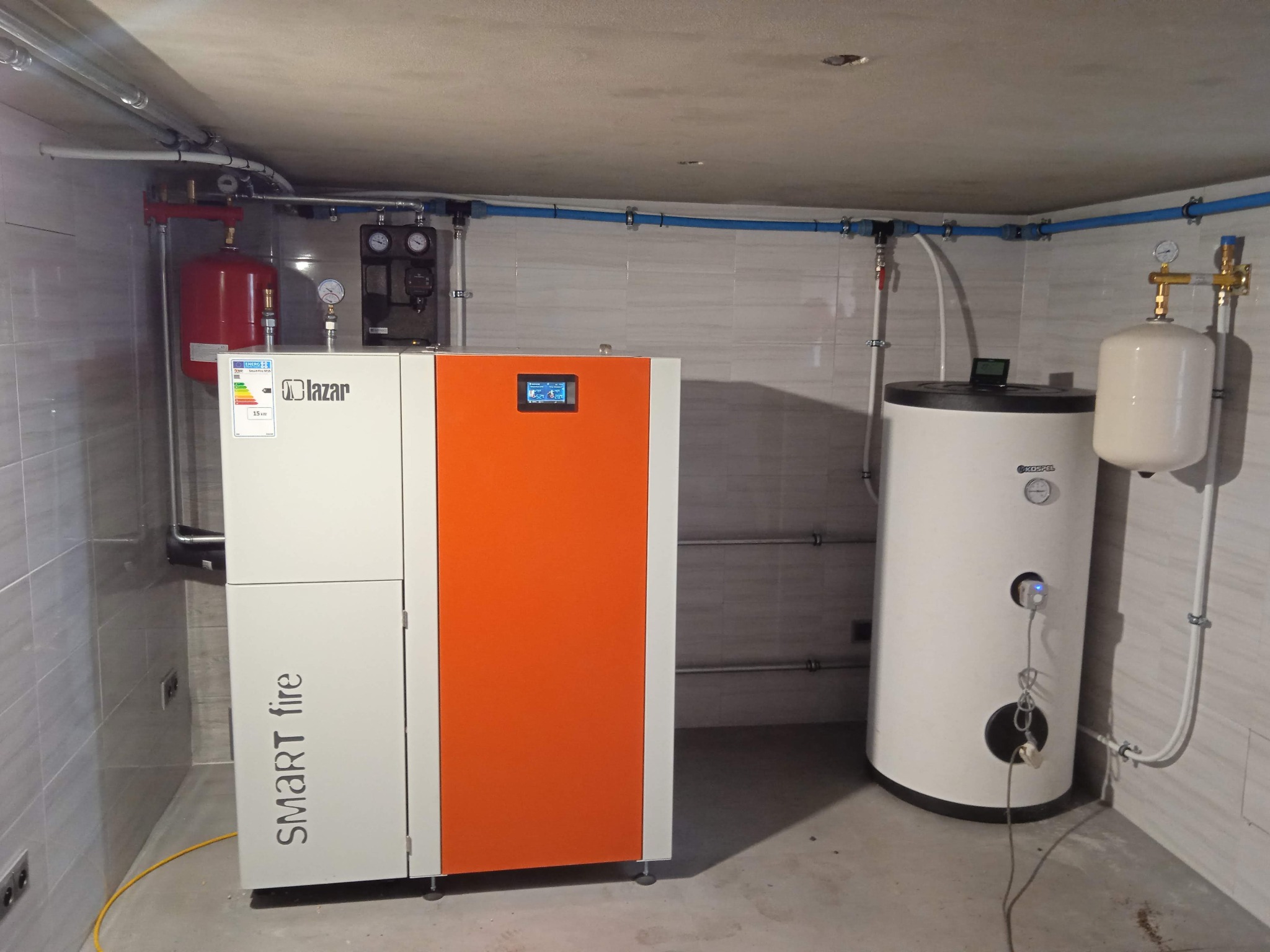 Nowoczesny, biało-pomarańczowy piec z podajnikiem marki Lazar Smart Fire, zbiornik akumulacyjny Kospel i instalacja CO w kotłowni wyłożonej jasnymi płytkami.