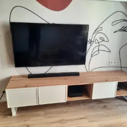 Nowoczesna szafka RTV z blatem drewnianym i białymi frontami, na tle ściany z abstrakcyjnym malunkiem, widoczne elementy wyposażenia wnętrza.