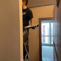 Szymon Ziółkowski - Mężczyzna na drabinie, w ubraniu roboczym z zabrudzeniami po gładzi, trzymający szpachelkę z gładzią, przygotowuje się do nałożenia jej na ścianę; zabezpieczone taśmą malarską obramowanie drzwi...