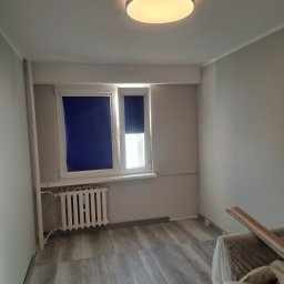 Szymon Ziółkowski - Pokój po remoncie z szarymi ścianami, panelami podłogowymi imitującymi drewno, białym grzejnikiem pod oknem z granatowymi roletami i okrągłą lampą sufitową. Część mebli zakryta folią.