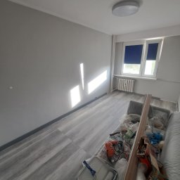 Szymon Ziółkowski - Pokój po malowaniu na szaro, z widocznymi narzędziami malarskimi i resztkami materiałów zabezpieczonych folią na kanapie, promienie słoneczne padają na świeżo pomalowaną ścianę.