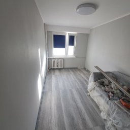 Szymon Ziółkowski - Pokój po świeżym malowaniu na szaro, z nową podłogą imitującą drewno, okno z zasłoniętą roletą i zakryty folią mebel z narzędziami malarskimi.