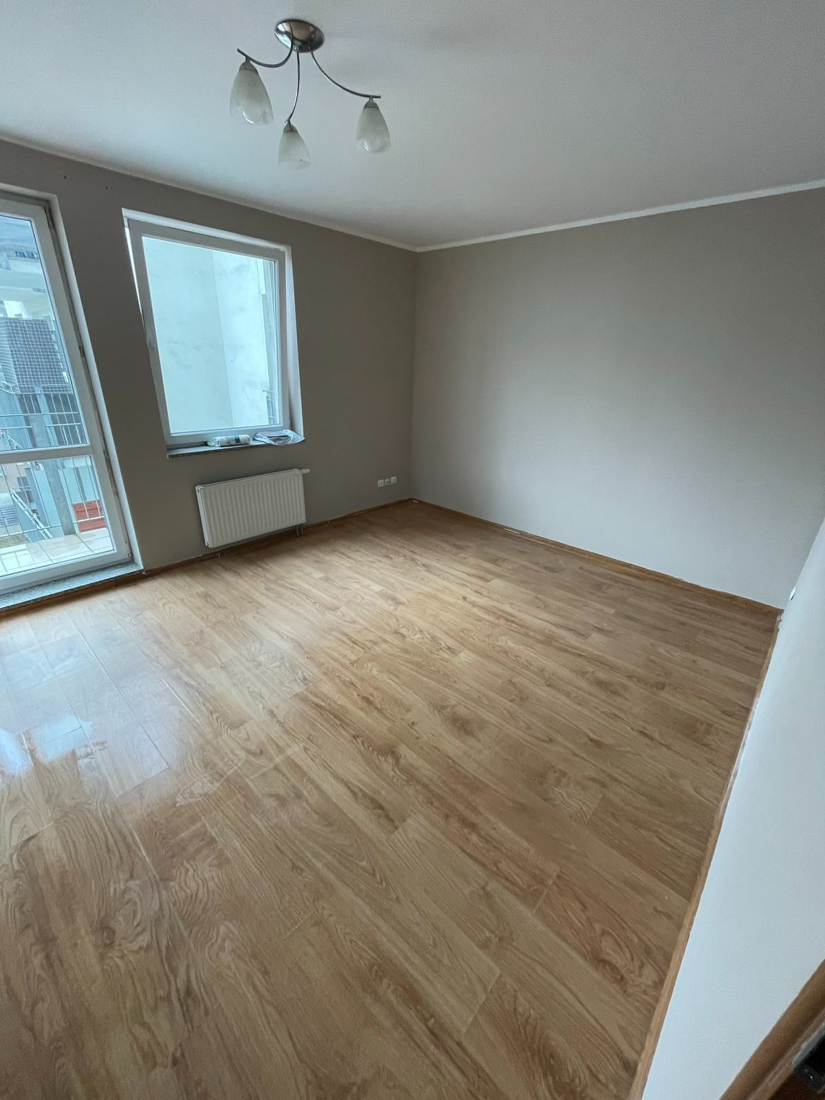 Pusty pokój po remoncie z nową podłogą z paneli imitujących drewno, świeżo pomalowanymi ścianami w odcieniach szarości i grzejnikiem pod oknem.