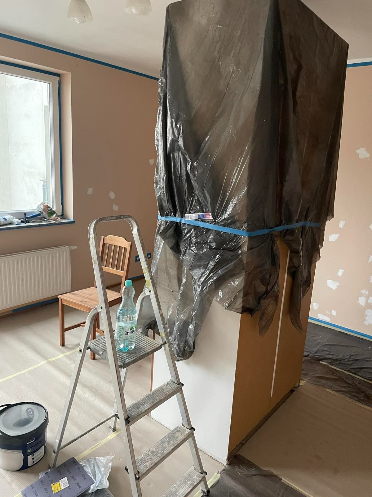 Pokój w trakcie przygotowań do malowania: meble zabezpieczone folią, ściany z widocznymi łatami po szpachlowaniu, taśma malarska przy suficie i oknach, drabina z butelką wody, wiadro z farbą...