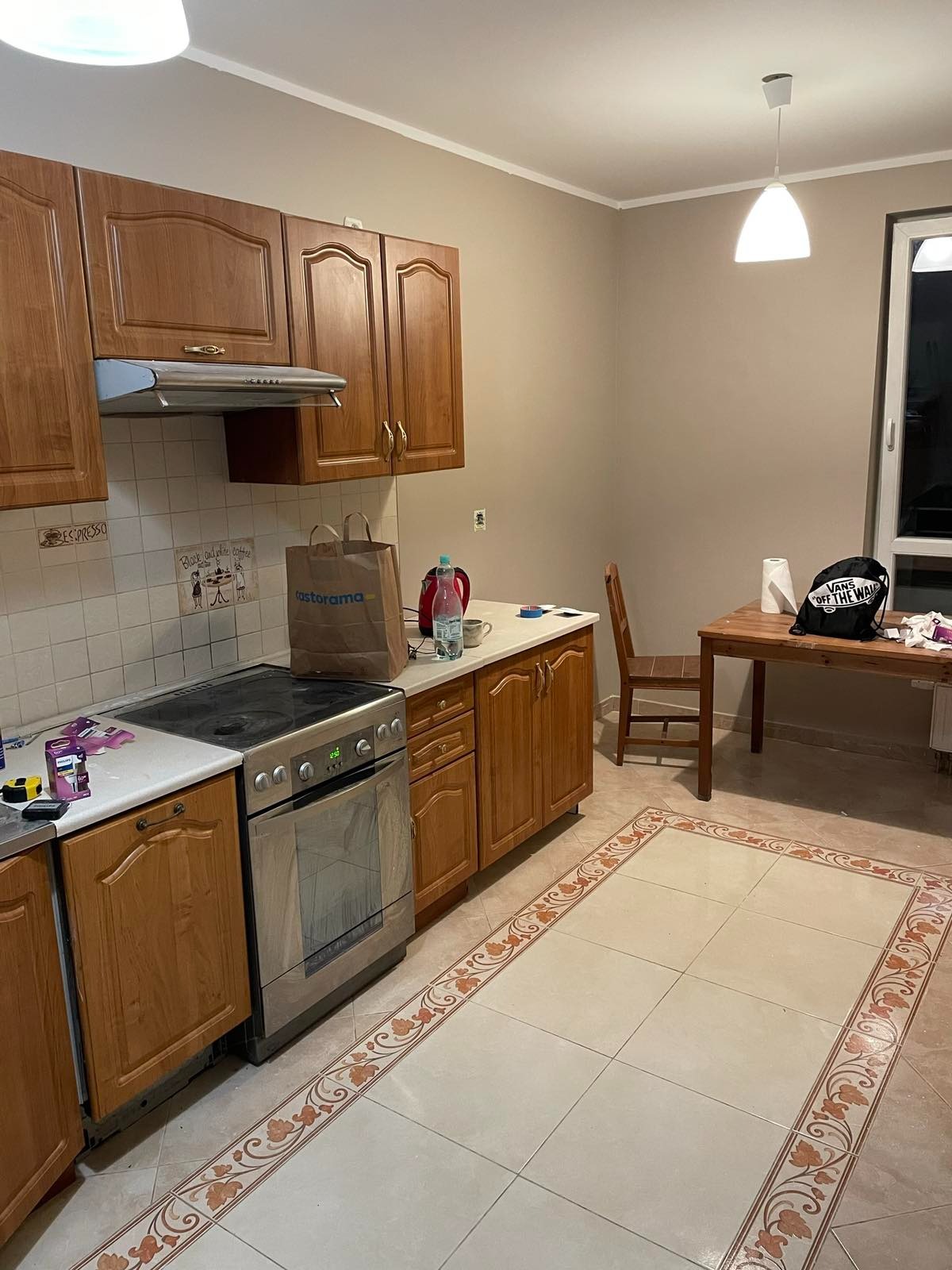 Kuchnia z drewnianymi szafkami, kuchenką elektryczną, beżowymi płytkami podłogowymi z dekoracyjnym obramowaniem, stołem i krzesłem, torbą Castorama na blacie oraz plecakiem Vans na stole przy oknie.