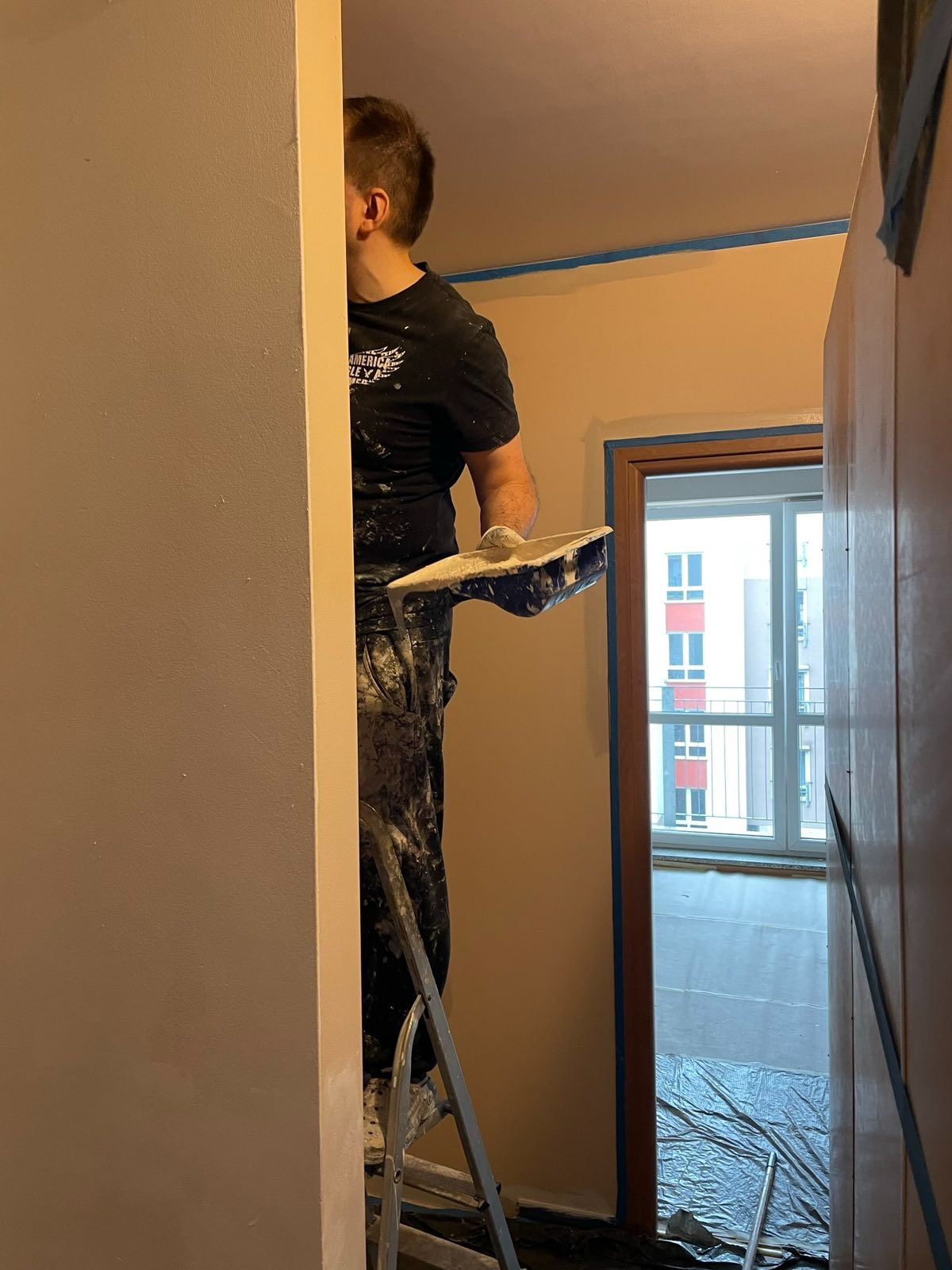 Mężczyzna na drabinie, w ubraniu roboczym z zabrudzeniami po gładzi, trzymający szpachelkę z gładzią, przygotowuje się do nałożenia jej na ścianę; zabezpieczone taśmą malarską obramowanie drzwi...