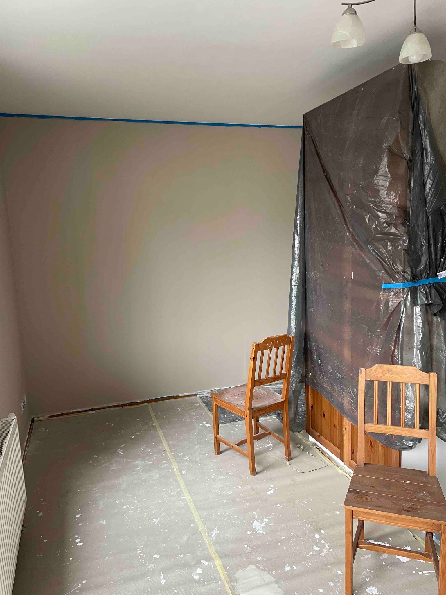 Pokój w trakcie prac przygotowawczych do malowania ścian: zabezpieczona folią meblościanka, dwie drewniane krzesła, podłoga zakryta folią malarską z widocznymi plamami farby, ściany z widocznym...