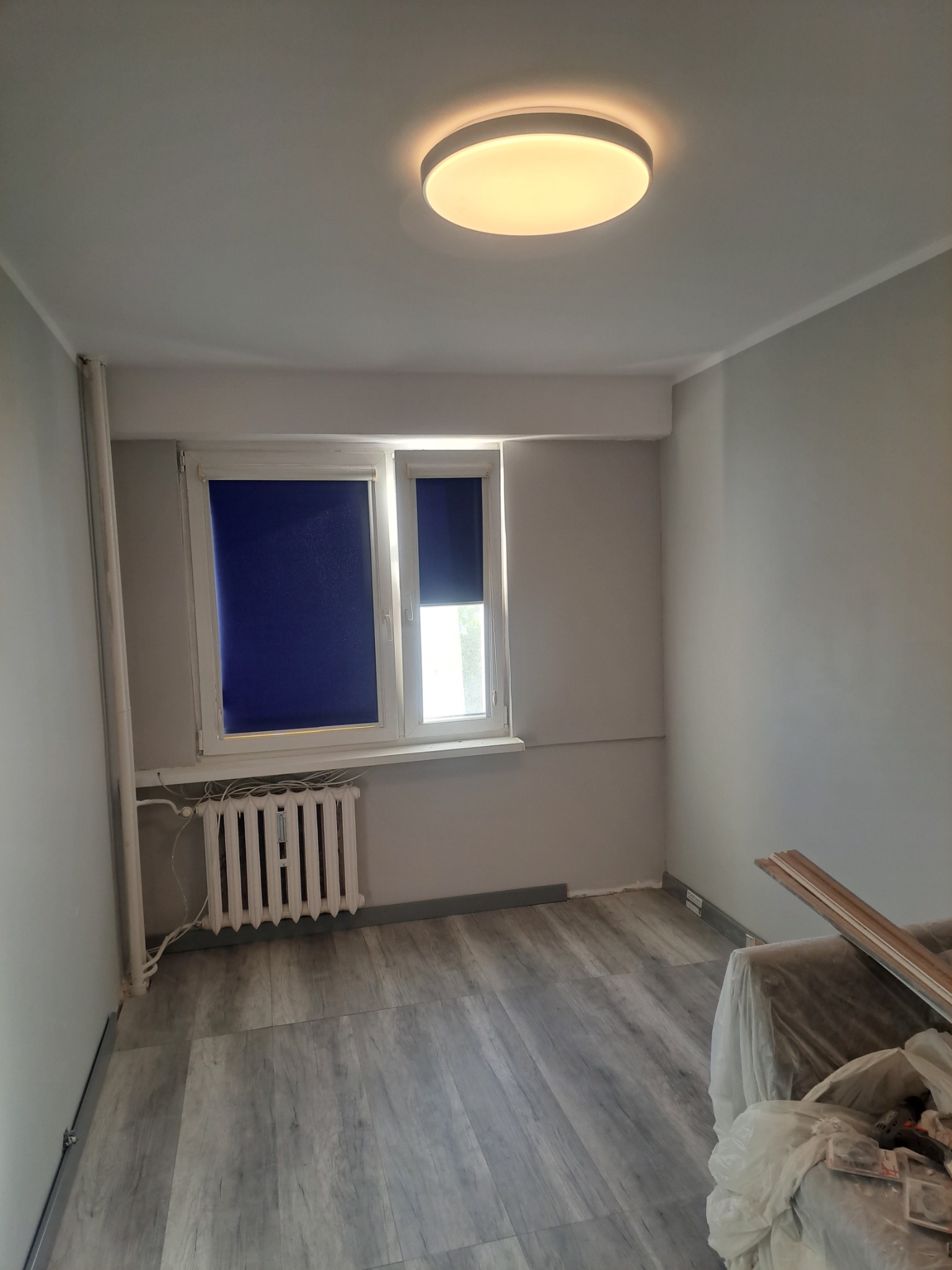 Pokój po remoncie z szarymi ścianami, panelami podłogowymi imitującymi drewno, białym grzejnikiem pod oknem z granatowymi roletami i okrągłą lampą sufitową. Część mebli zakryta folią.