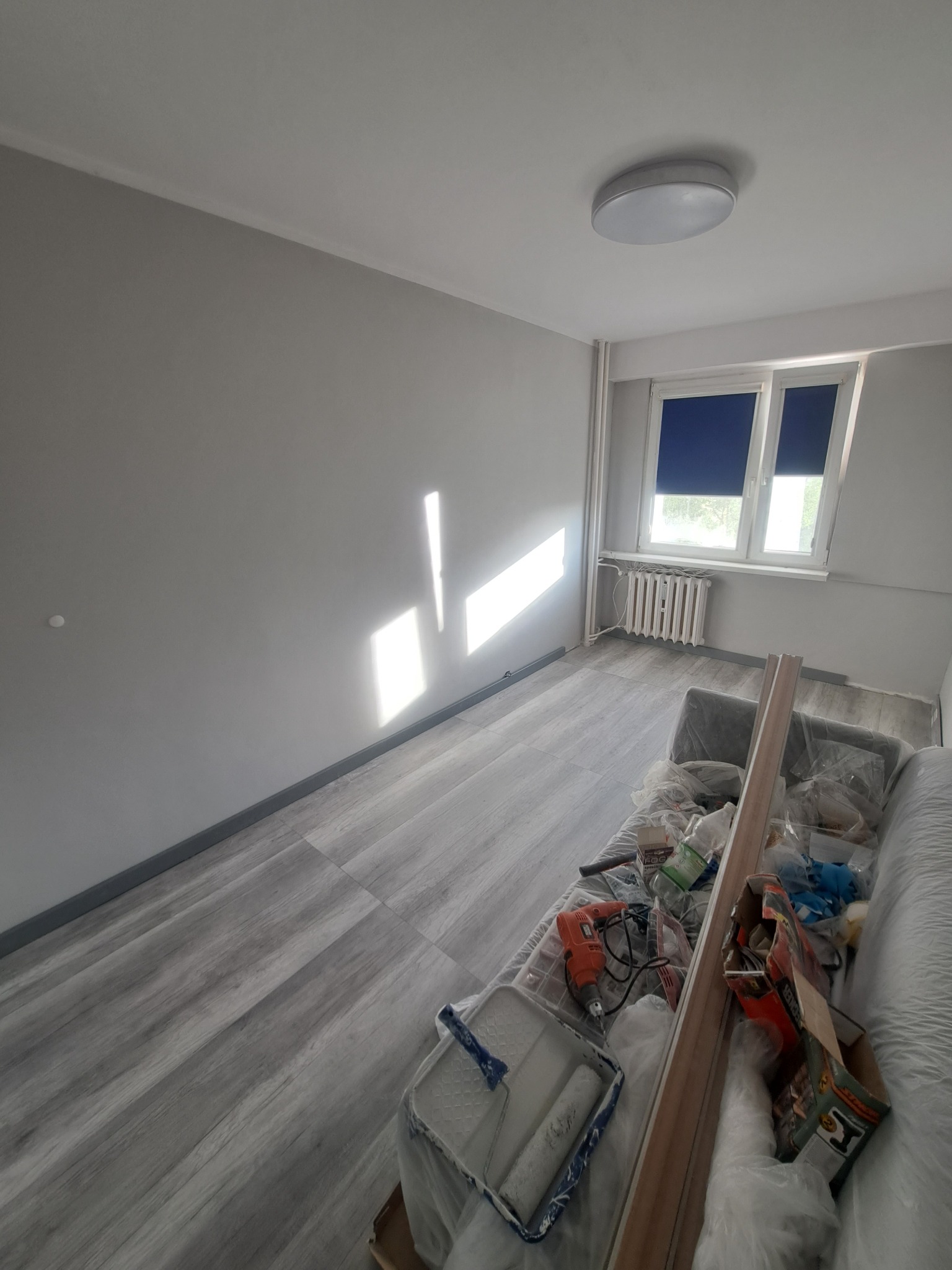 Pokój po malowaniu na szaro, z widocznymi narzędziami malarskimi i resztkami materiałów zabezpieczonych folią na kanapie, promienie słoneczne padają na świeżo pomalowaną ścianę.
